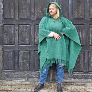 K&ouml;nnte beinhalten: Ein gr&uuml;ner Kapuzenponcho mit Fransen. Der Poncho hat einen Knopfverschluss am Halsausschnitt und ist aus einem weichen, strukturierten Stoff gefertigt. Das Model tr&auml;gt den Poncho mit blauen Jeans und schwarzen Stiefeln.