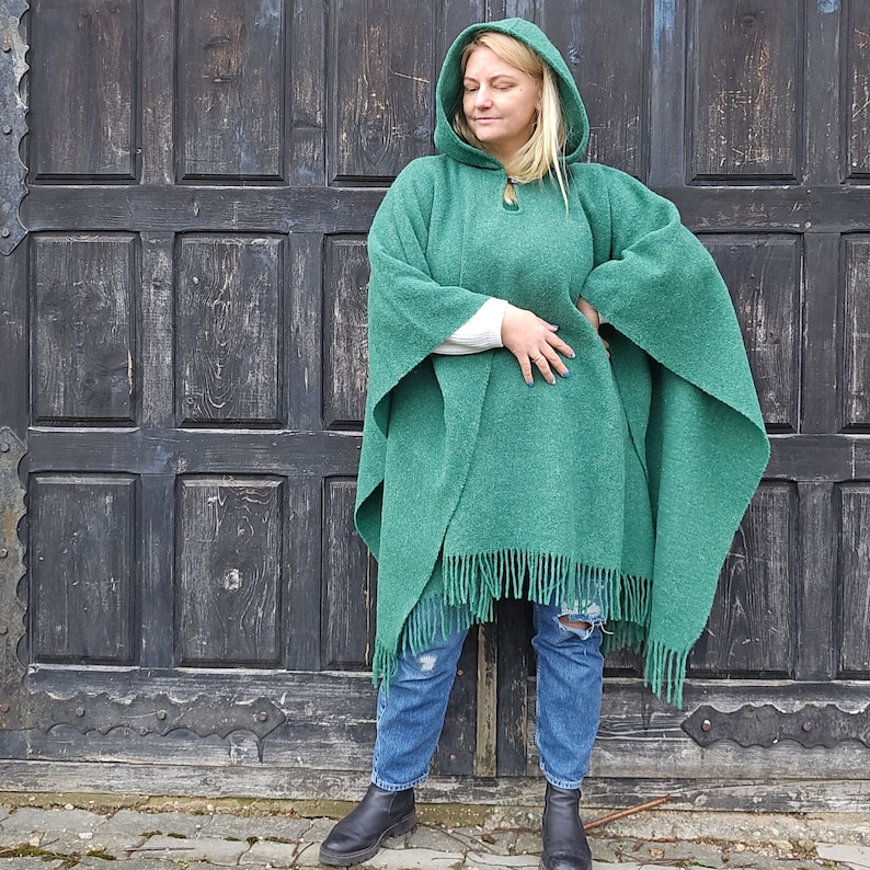 K&ouml;nnte beinhalten: Ein gr&uuml;ner Kapuzenponcho mit Fransen. Der Poncho hat einen Knopfverschluss am Halsausschnitt und ist aus einem weichen, strukturierten Stoff gefertigt. Das Model tr&auml;gt den Poncho mit blauen Jeans und schwarzen Stiefeln.