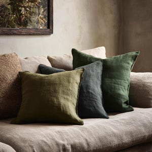 Può includere: Una collezione di cuscini quadrati in varie tonalità di verde e grigio, disposti su un divano dai toni neutri. I cuscini hanno un aspetto strutturato, simile alla lino, creando un'atmosfera accogliente e invitante. Un'immagine incorniciata è sullo sfondo.