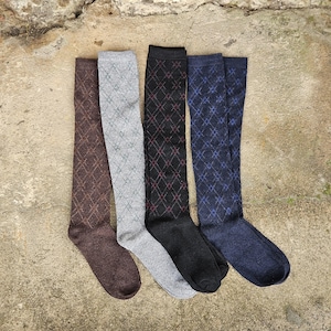 Può includere: Quattro paia di calze al ginocchio in vari colori: marrone, grigio, nero e blu navy. Ogni calza presenta un motivo a rombi in un colore a contrasto. Le calze sono disposte su una superficie strutturata dai toni neutri.