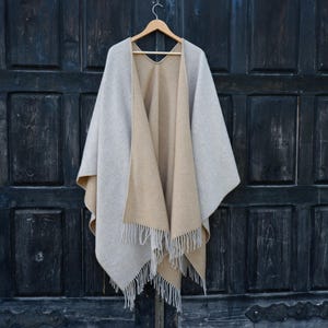 May include: A reversible cape in light gray and tan, hanging on a wooden hanger. The cape has a fringed hemline and is displayed against a dark wooden door. The garment appears to be made of a soft, woven material.