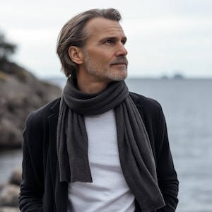 May include: A dark gray scarf draped over a white t-shirt and black jacket. The scarf is made of a soft material and is wrapped around the neck. The man is looking off to the side with a thoughtful expression.