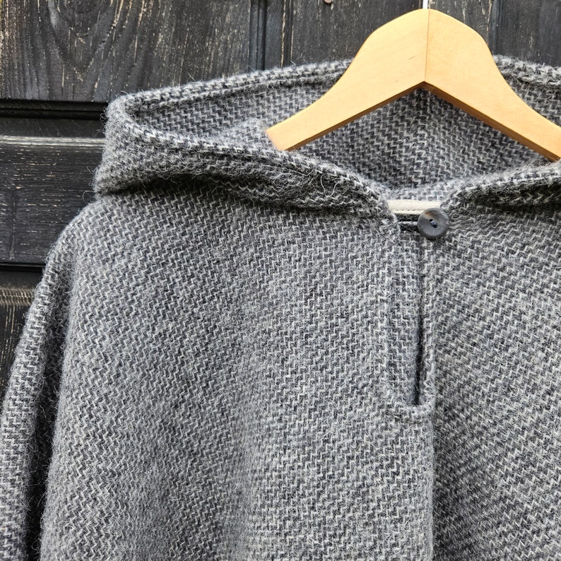 K&ouml;nnte beinhalten: Ein grauer Kapuzenponcho aus gewebtem Stoff, der an einem Holzkleiderb&uuml;gel h&auml;ngt. Der Poncho hat einen Knopfverschluss am Halsausschnitt. Der Stoff hat ein strukturiertes Aussehen mit einem dezenten Fischgr&auml;tmuster.