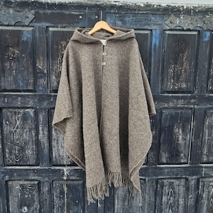 K&ouml;nnte beinhalten: Ein brauner Kapuzenponcho mit zwei Knopfverschl&uuml;ssen und Fransen am Saum. Der Poncho besteht aus einem strukturierten Stoff und h&auml;ngt an einem Holzb&uuml;gel vor einer dunklen Holzt&uuml;r.