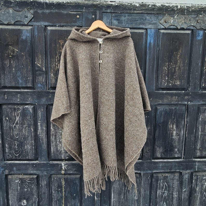 K&ouml;nnte beinhalten: Ein brauner Kapuzenponcho mit zwei Knopfverschl&uuml;ssen und Fransen am Saum. Der Poncho besteht aus einem strukturierten Stoff und h&auml;ngt an einem Holzb&uuml;gel vor einer dunklen Holzt&uuml;r.