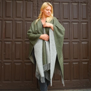 May include: A reversible cape in olive green and gray. The cape has a fringed edge on the gray side. The person is wearing the cape in front of a brown wooden door.