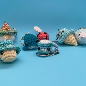 May include: A collection of crocheted keychains featuring various designs, including a mushroom, a bee, an ice cream cone, and a turtle. The keychains are made with colorful yarn and have cute, whimsical faces.