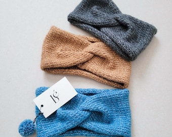 May include: Three knitted headbands in shades of blue, tan, and grey are displayed. The blue headband has a small pom-pom attached. A white tag with the letters "is" is attached to the blue headband. The headbands are made of a soft, textured yarn.