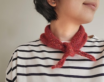 May include: A rust-colored, hand-knitted neckerchief featuring a textured, speckled yarn finish. The accessory is tied in a simple knot at the front of the neck, resting against a white shirt with horizontal black stripes.