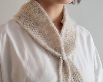 May include: A white knitted scarf with a delicate texture, tied in a loose knot around the neck with a small silver pin.