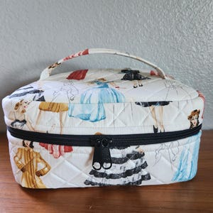 May include: A quilted cosmetic bag with a black zipper and handle. The bag is white with a repeating pattern of vintage fashion illustrations in various colors. The bag is sitting on a wooden surface.