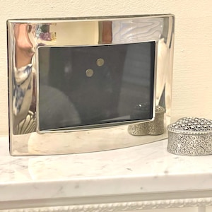 May include: A polished silver picture frame with a curved top, containing a black insert. A small, decorative silver box with an ornate design is positioned to the right. The frame and box are on a white surface.