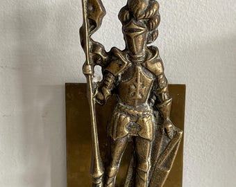 Op de afbeelding: Een koperen boekensteun met een staande ridder in volledig harnas, die een lange speer met een vlag in de rechterhand en een schild in de linkerhand vasthoudt. De ridder is gemonteerd op een platte, rechthoekige koperen basisplaat.