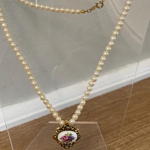 May include: A white pearl necklace with a gold pendant featuring a floral design. The pendant has a white porcelain centre with pink and purple flowers.