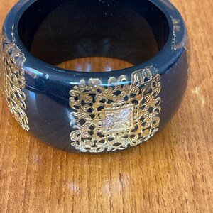Peut inclure: Un large bracelet jonc noir avec des motifs de filigrane dorés. Le bracelet a une finition brillante et une forme arrondie. Les motifs dorés sont symétriques et ornés, ajoutant une touche d'élégance à l'accessoire.