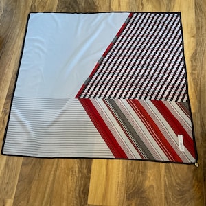 Peut inclure: Un foulard carré avec un motif géométrique. Le foulard présente une section bleu clair, une section avec des rayures diagonales noires et blanches et une section avec des rayures rouges et grises. Le foulard a une bordure noire et le texte "OREY CYRECO".