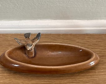 Könnte beinhalten: Eine braune Keramikschale mit einer kleinen Vogelfigur darauf. Die Schale ist oval geformt und hat eine glänzende Oberfläche.