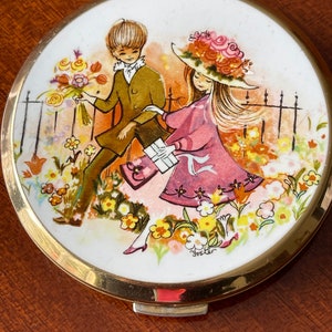 以下が含まれることがあります： 金色の縁取りと白い磁器の蓋が付いたヴィンテージのコンパクトミラー。蓋には、花とフェンスのある庭園で、男の子と女の子が描かれたカラフルなイラストが描かれています。蓋の下部には「Foster」という文字が印刷されています。