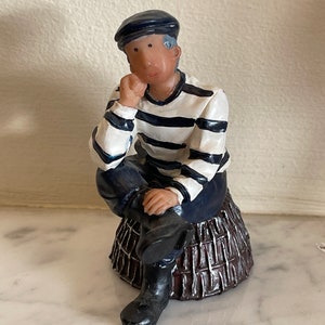 Peut inclure: Figurine d'une personne en tenue de marin, assise sur un panier marron. La figurine porte un béret bleu marine, une chemise rayée blanche et marine, un pantalon bleu marine et des bottes noires. La personne se repose le menton sur la main.