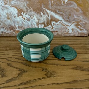 May include: A small green and white ceramic jar with a lid. The jar has a plaid pattern and the lid has a small knob on top.