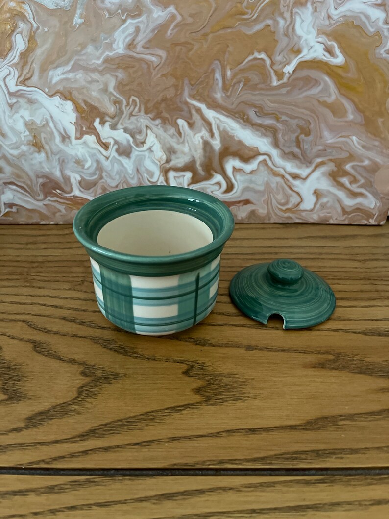 May include: A small green and white ceramic jar with a lid. The jar has a plaid pattern and the lid has a small knob on top.