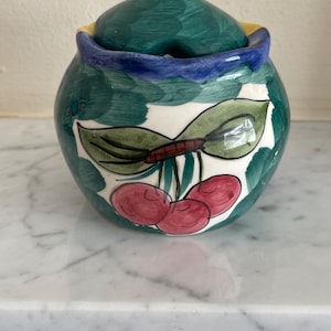 Puede incluir: Un azucarero de cerámica pintado a mano con tapa. El cuenco es redondo con un fondo verde y blanco y un borde azul. El cuenco presenta un diseño pintado de cerezas y hojas. La tapa tiene un pomo amarillo.