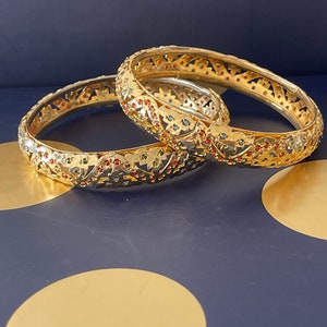 May include: Two gold bangles with intricate designs and colourful gemstones. The bangles are stacked on top of each other.