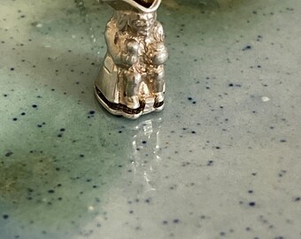 May include: A silver charm necklace featuring a detailed figurine of a seated man wearing a hat. The charm is in the shape of a pitcher.