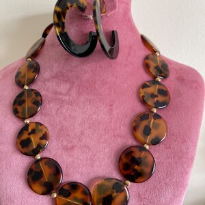 May include: A brown and black tortoiseshell necklace with large round beads and gold accents. The necklace is on a pink mannequin.
