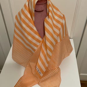 May include: An orange and white striped scarf with the brand name "LEXON" visible. The scarf is draped over a pink form. The scarf has a repeating pattern of orange and white stripes. The scarf is made of a lightweight material.