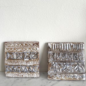 May include: Two square ceramic tiles featuring a brown base with white glazed relief patterns. The left tile displays horizontal bands of waves, squares with crosses, vertical lines, and triangles. The right tile shows vertical lines, squares with crosses, vertical dashes, and triangular shapes.