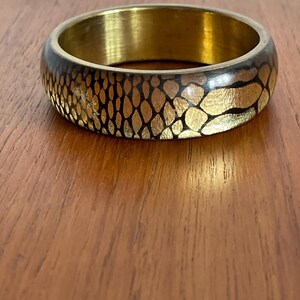 May include: A gold-toned bangle bracelet with a dark brown exterior featuring a gold-colored scale pattern. The interior of the bracelet is a solid gold color. The bracelet is made of metal and is a circular shape.