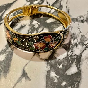 May include: A gold bangle bracelet with a black enamel design featuring colourful flowers and butterflies. The bracelet has a hinged clasp.