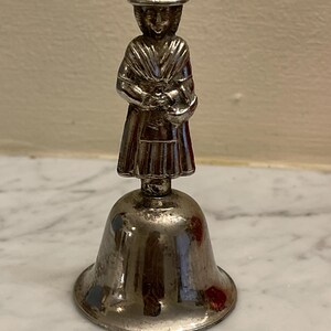 Op de afbeelding: Zilveren bel met een beeldje van een vrouw die een hoed en een lange jurk draagt. De vrouw houdt een mand in haar handen.