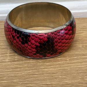 May include: A wide, round bangle bracelet with a snakeskin pattern in shades of red and black. The bracelet has a silver-colored metal trim and a gold-colored interior.