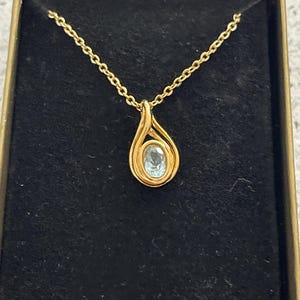 May include: A gold necklace with a teardrop-shaped pendant featuring a light blue gemstone.