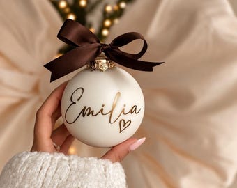 Könnte beinhalten: Eine runde weiße Weihnachtskugel mit dem Namen Emilia in eleganter goldener Schreibschrift und einem kleinen goldenen Herz darunter. Die Kugel ist mit einer dunkelbraunen Satinschleife am metallenen Aufhänger verziert.