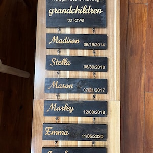 May include: A wooden sign with the text "Happiness is having grandchildren to love" and a series of smaller signs with the names of grandchildren and their birth dates. The names are Madison, Stella, Mason, Marley, Emma, and Jacob.