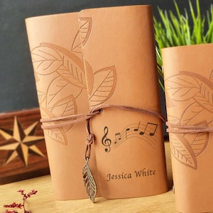 May include: A brown leather journal with a leaf design and a music note and treble clef design. The journal has a brown leather strap and a feather charm. The journal is personalised with the name "Jessica White".