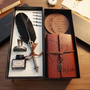 Könnte beinhalten: Ein Vintage-Schreibset in einer schwarzen Box. Es enthält eine schwarze Federkiel, Tinte, Federn und ein ledergebundenes Notizbuch. Ein Holzuntersetzer mit graviertem Zitat und eine kleine Uhr sind ebenfalls zu sehen.