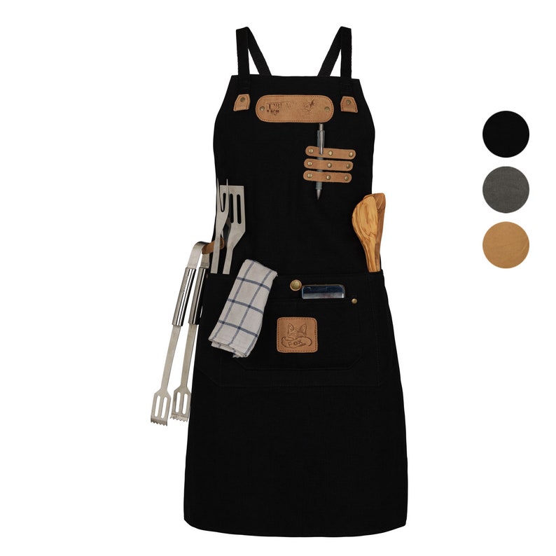 May include: Black canvas apron with leather straps and pockets. The apron has a brown leather patch with the text "The Fox & The Flame" and a small pocket with a metal tool holder. The apron is shown with a wooden spoon and metal grilling tools.