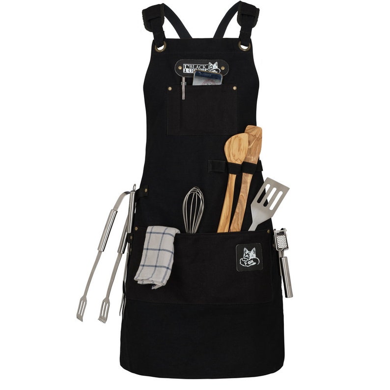 May include: Black canvas apron with multiple pockets and a black and white fox logo. The apron has a variety of kitchen tools in the pockets, including a whisk, wooden spoons, tongs, and a garlic press.