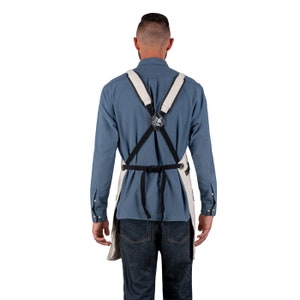 May include: A person wearing a white canvas apron with adjustable black straps and a black buckle. The apron has a black and white logo on the back.