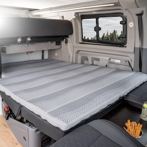 Könnte beinhalten: Innenansicht eines Wohnmobils mit einer grau-weiß gestreiften Matratze. Das Bett ist gemacht, und ein Fenster zeigt eine Aussicht auf Bäume. Der Van hat graue Sitze und einen schwarzen Stauraum über Kopf.