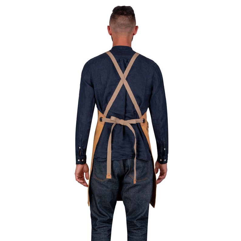May include: A person wearing a blue shirt and jeans with a tan apron tied around their waist. The apron has adjustable straps that cross in the back.