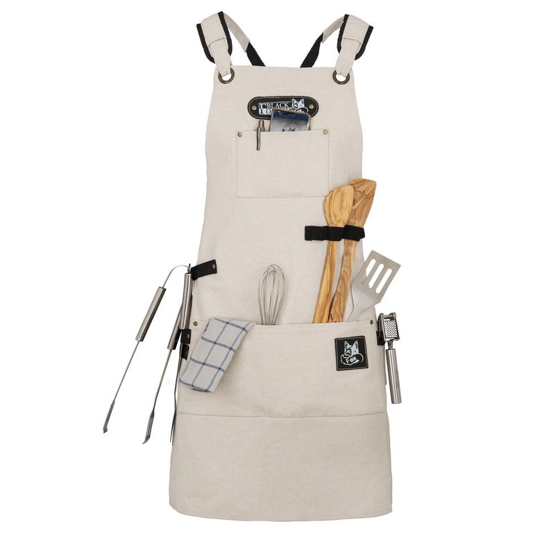 May include: A beige canvas apron with adjustable straps, a large pocket, and a smaller pocket with a leather patch that says "Black Fox Goods". The apron has a variety of tools hanging from it, including a whisk, wooden spoons, a spatula, tongs, and a garlic press.