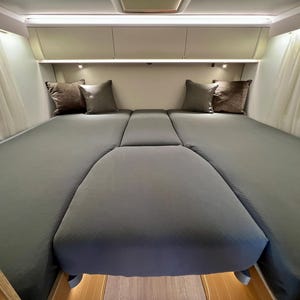 Może przedstawiać: Szare, L-kształtne łóżko z dwoma poduszkami po obu stronach. Łóżko jest pościelone szarą kołdrą i znajduje się w kamperze.