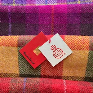 May include: Close-up of colorful Harris Tweed fabric in various plaid patterns. The colors include shades of purple, pink, red, orange, and green. Two tags are visible, one red with the text "Harris Tweed" and another white with a logo.