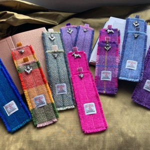 May include: A selection of tweed bookmarks in various colours, including blue, pink, purple, and green. Each bookmark features a decorative metal charm and a small tag with text. The bookmarks are displayed on open books, showcasing their texture.