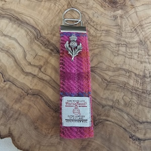 May include: A pink and blue tweed keychain with a silver thistle charm and a white label that reads "HAND WOVEN CLOTH Harris Tweed OUTER HEBRIDES OF SCOTLAND 100% PURE NEW WOOL"